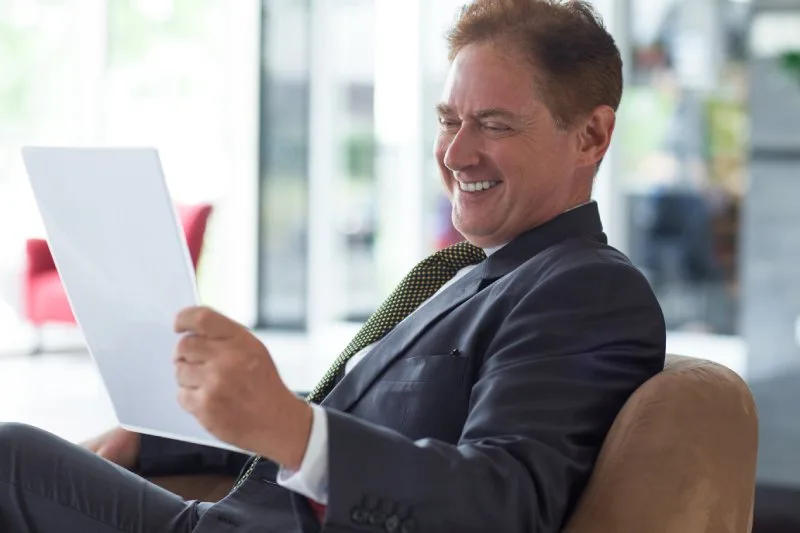 Portrait of positive senior Caucasian businessman wearing suit sitting on coach reading document and smiling